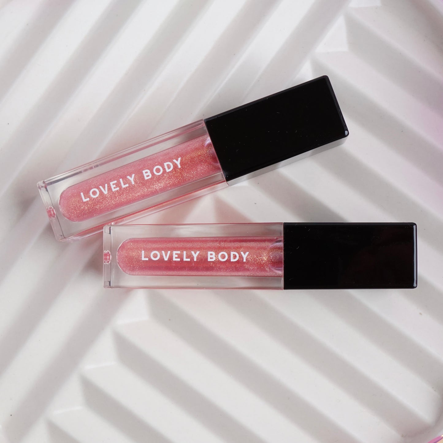 Two 'Lovely Body' lip glosses on a white textured background