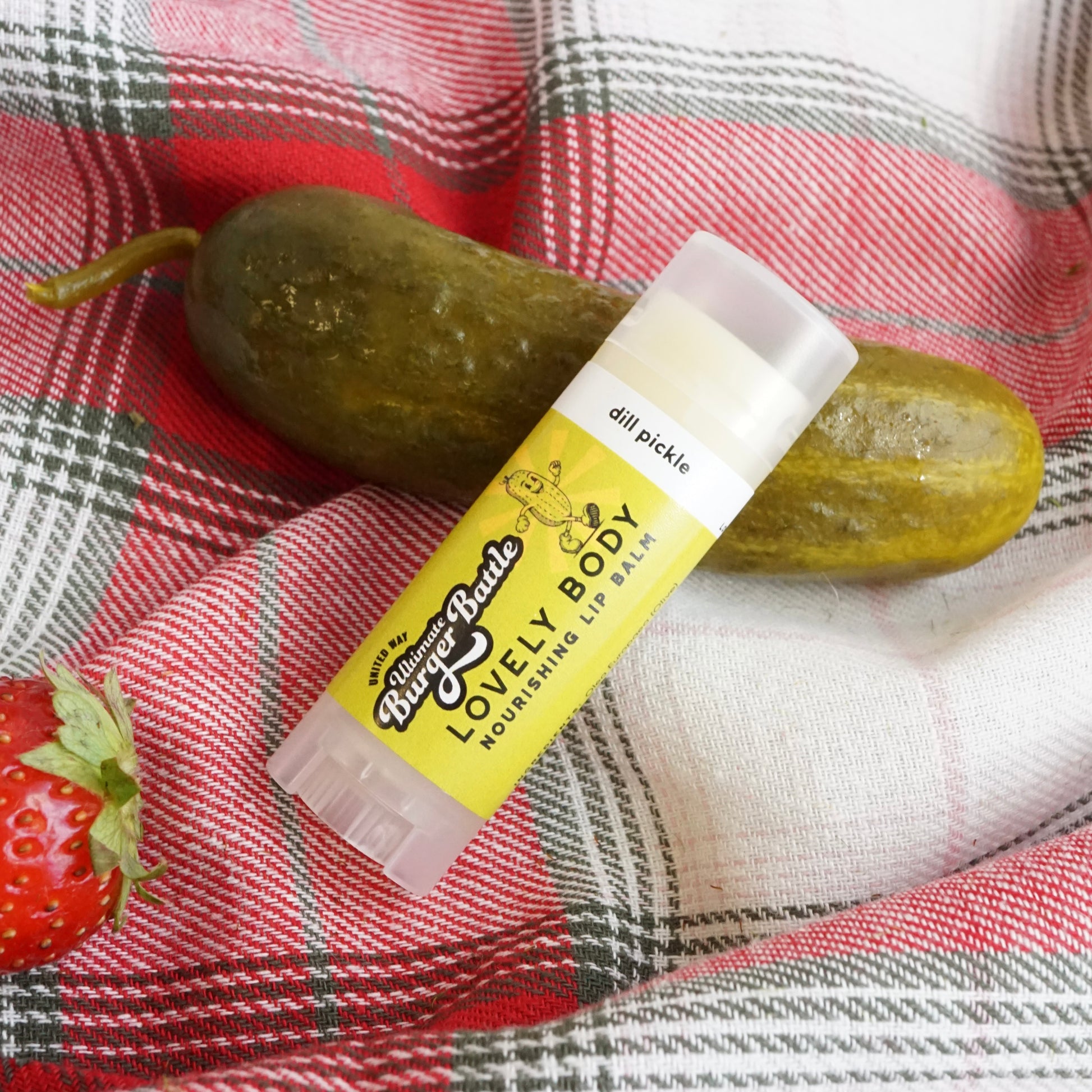 Lip balm labeled 'Lovely Body' with a pickle and strawberry on a plaid background