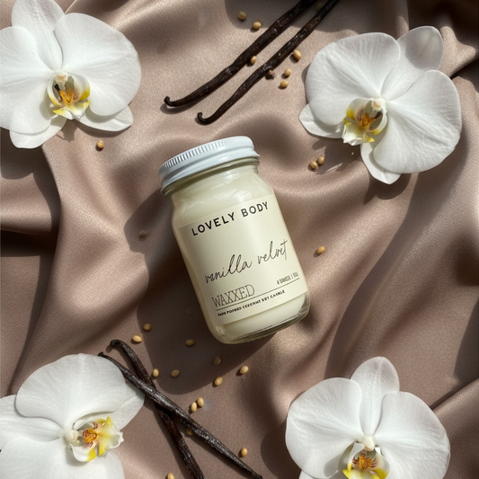 A glass jar candle labeled 'LOVING BODY vanilla velvet' sits on draped taupe satin fabric, surrounded by white orchids, vanilla beans, and small seeds. The candle has a white label and a silver metal lid.