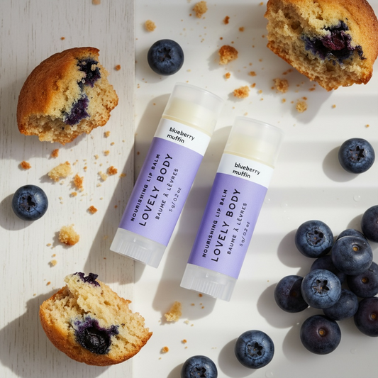 Two lip balm tubes labeled 'Lovely Body Nourishing Lip Balm' in blueberry muffin scent, styled on a white surface with blueberries, crumbs, and half-muffins.