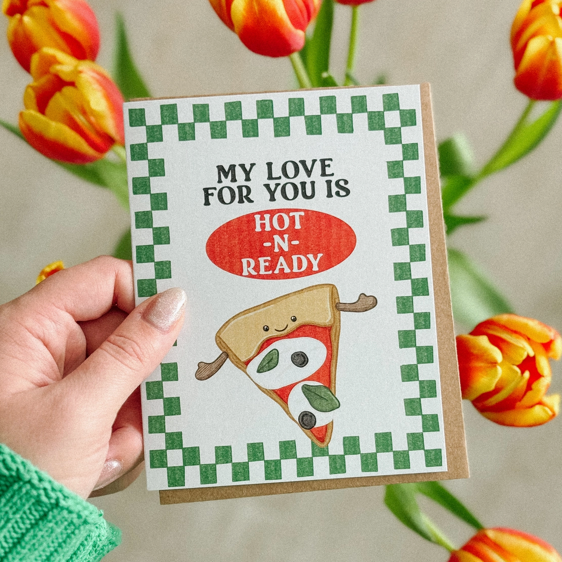 Card with pizza slice illustration and text held by a hand, with tulips in the background