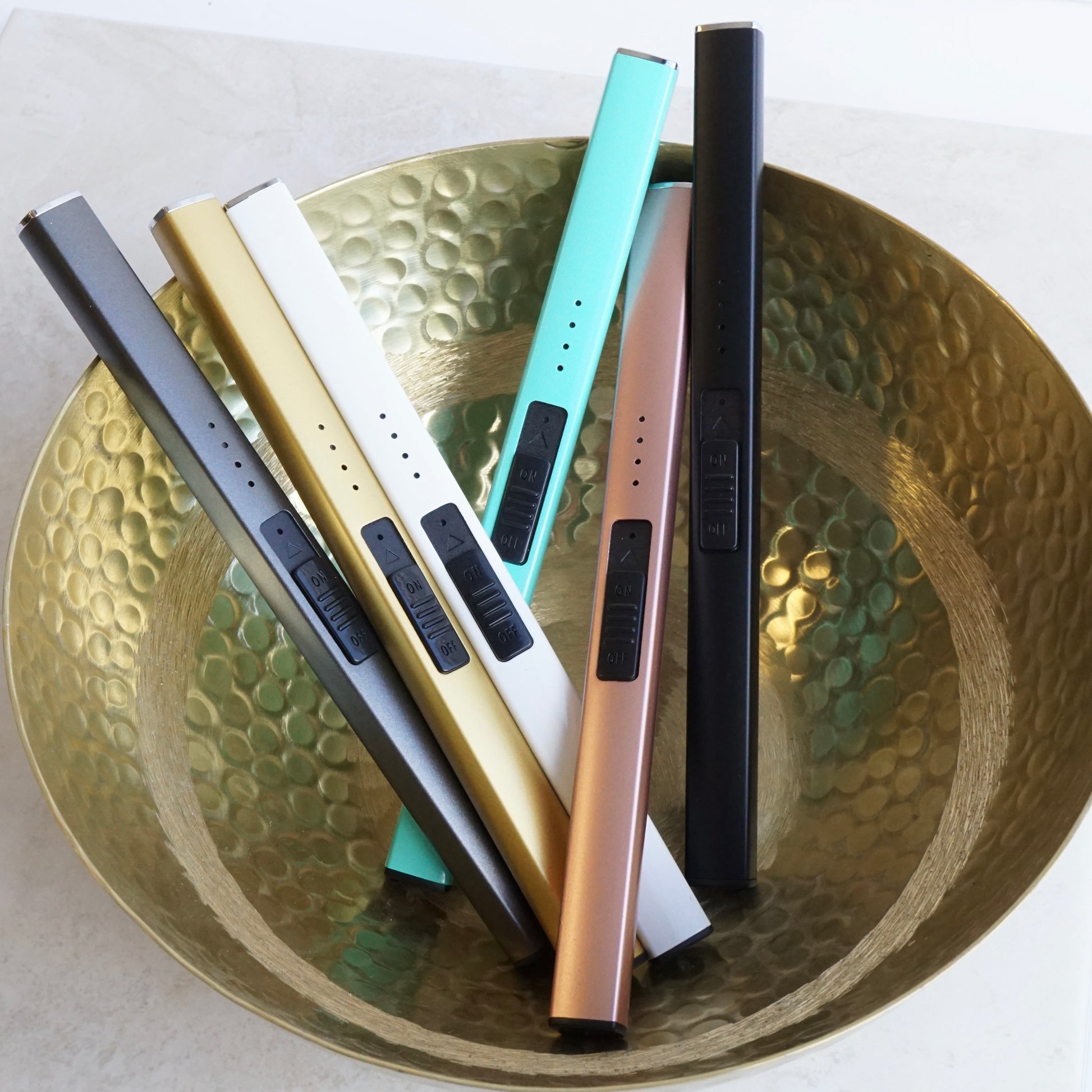 A gold bowl displaying 6 different coloured lighters, grey, gold, white, teal, rose gold, black