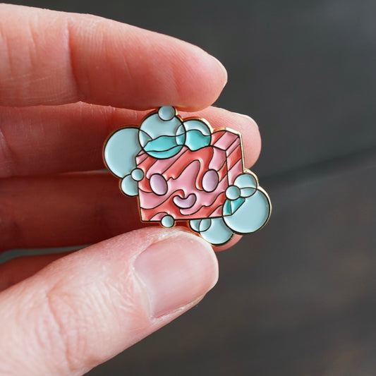 Small enamel pin of a cartoon bar of soap, in pinks and blues.